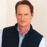 A person with short brown hair is smiling while wearing a light blue shirt and dark vest, standing against a light background.