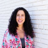 Person with curly hair smiling in front of a white brick wall, wearing a floral-patterned top.