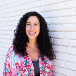 Person with curly hair smiling in front of a white brick wall, wearing a floral-patterned top.