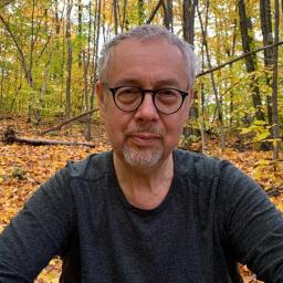A person with glasses and a gray beard is in a forest with autumn leaves on the ground. They are wearing a gray shirt.