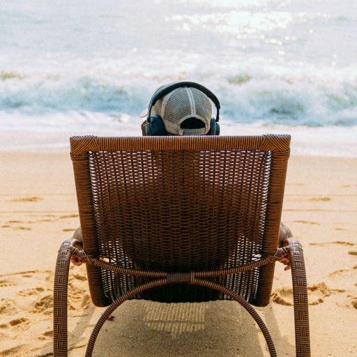 Person wearing headphones sits in a wicker chair facing the ocean on a sandy beach, with waves in the background and footprints in the sand.