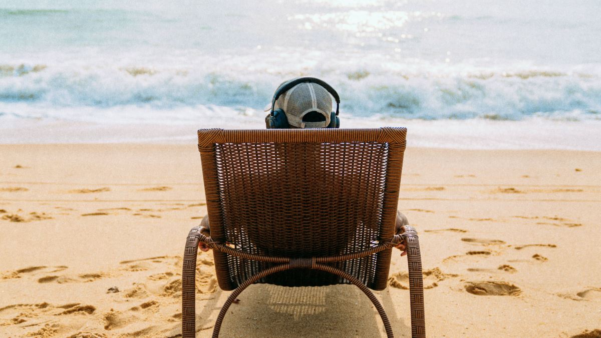 Person wearing headphones sits in a wicker chair facing the ocean on a sandy beach, with waves in the background and footprints in the sand.