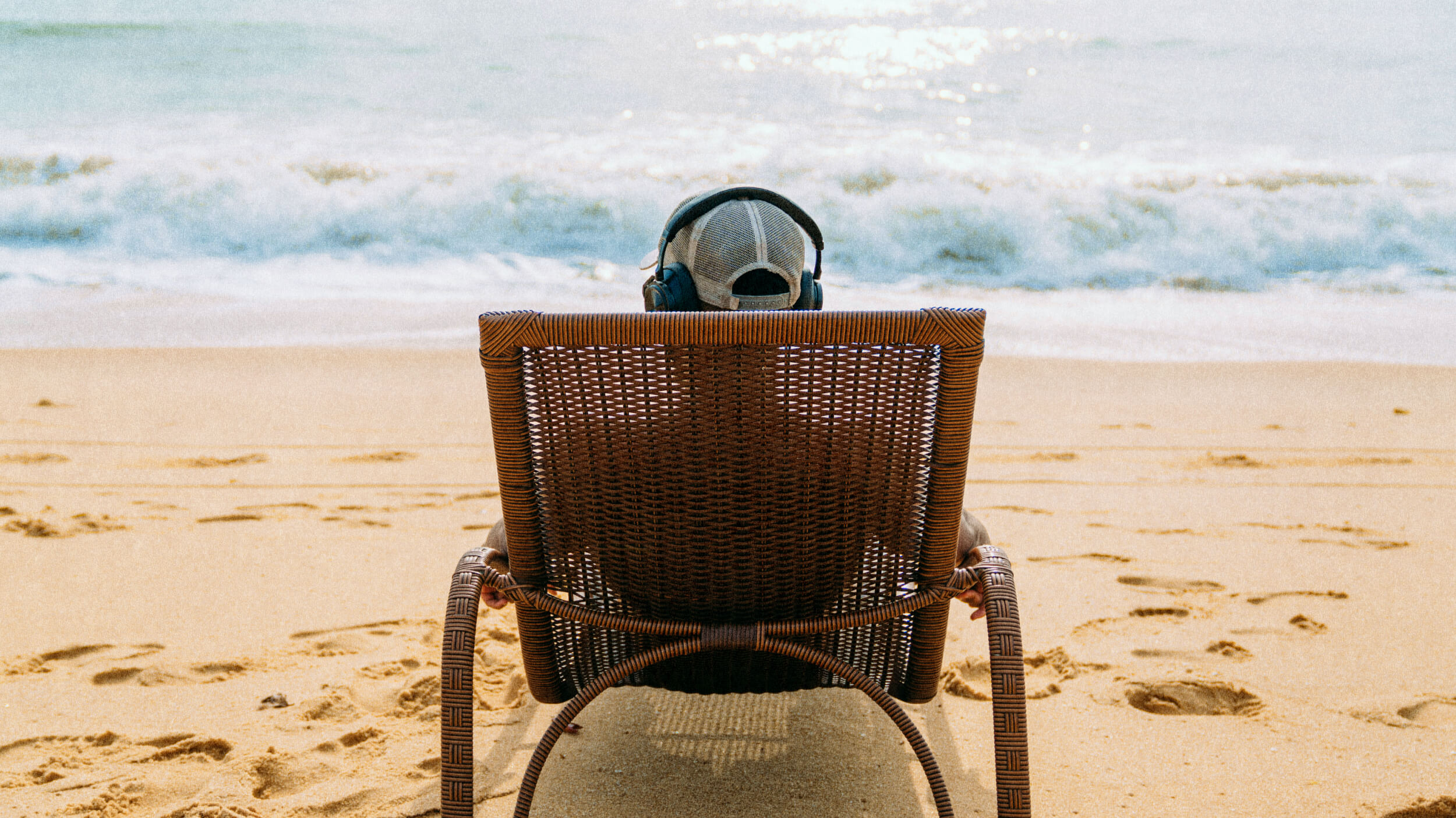 Person wearing headphones sits in a wicker chair facing the ocean on a sandy beach, with waves in the background and footprints in the sand.