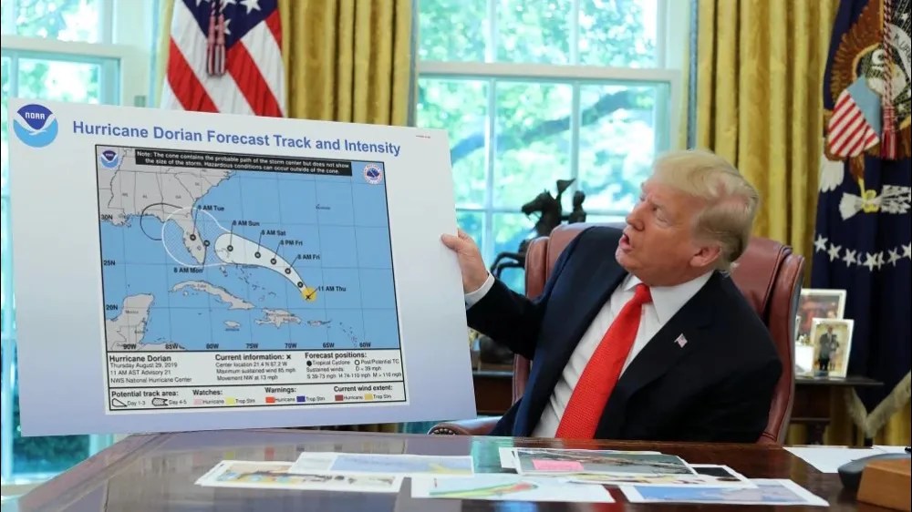 A man in a suit sits at a desk holding a large Hurricane Dorian forecast map, with U.S. flags and official emblems visible in the room, evoking the urgency of a 1938 science manifesto warning about threats to democracy.