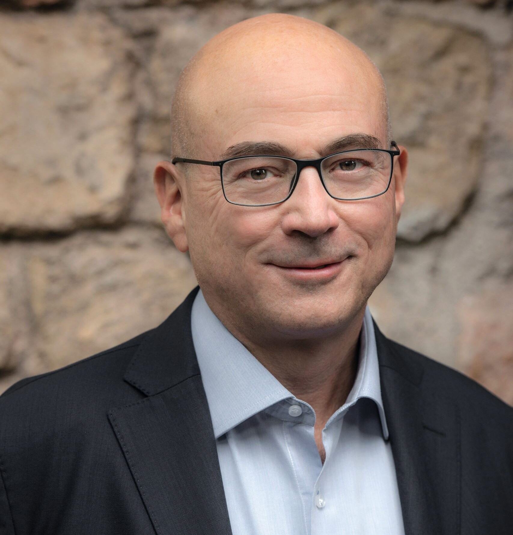A bald man wearing glasses, a light blue shirt, and a dark blazer is standing in front of a stone wall.