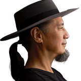 A man with a black ponytail and goatee wearing a wide-brimmed hat and black shirt is shown in profile against a dark background.