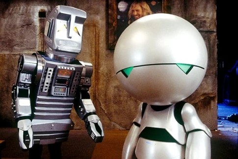 Two humanoid robots stand side by side in a dimly lit room with a weathered wall and a framed picture in the background.