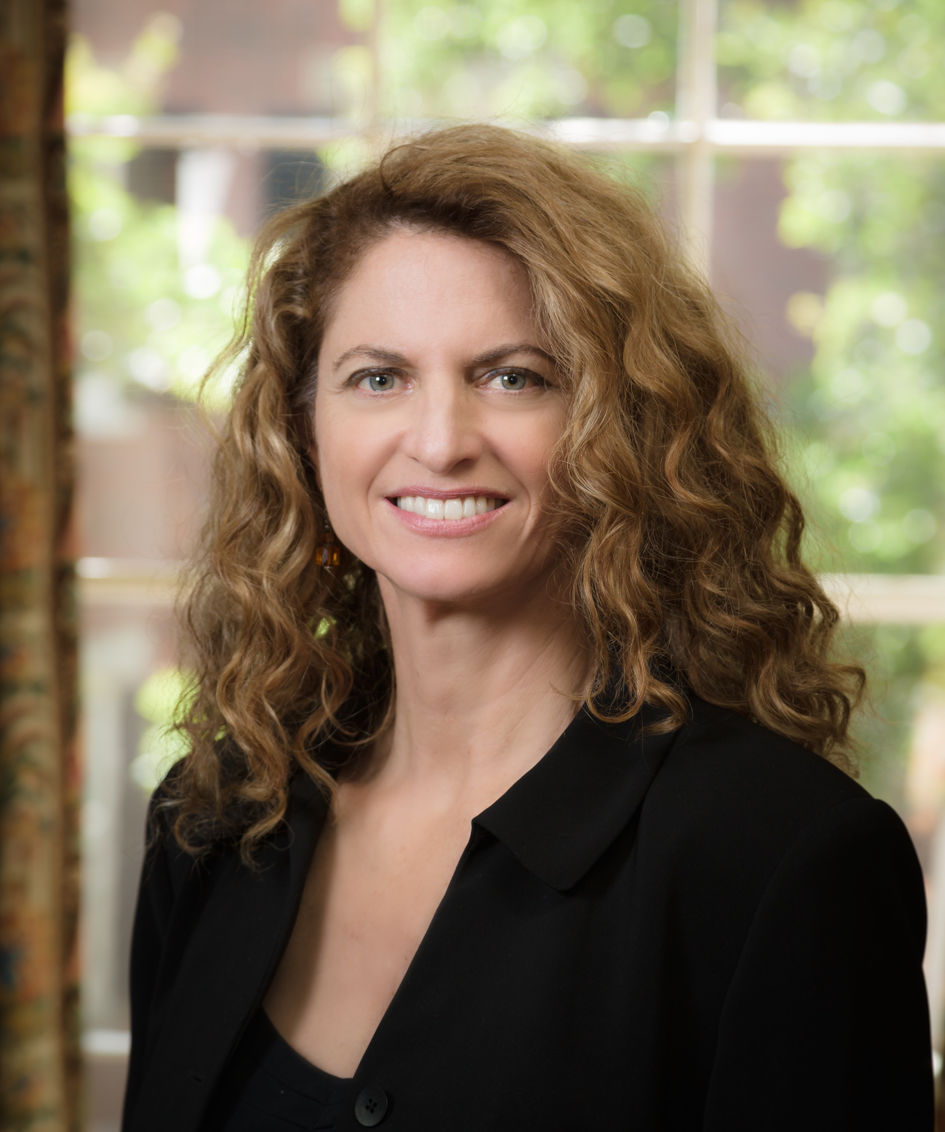 A woman with curly light brown hair, wearing a black blazer, stands indoors in front of a window with greenery visible outside.