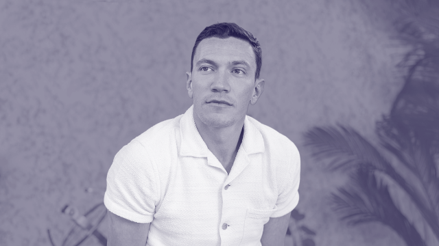 David Perell, in a short-sleeve white shirt, sits in front of a blurred background with plants, looking slightly to the side.