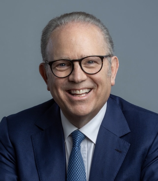 A middle-aged man with gray hair and glasses smiles while wearing a blue suit, white shirt, and blue patterned tie against a plain gray background.