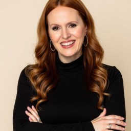 Woman with long red hair in loose curls, wearing a black turtleneck and hoop earrings, smiling with arms crossed against a beige background.