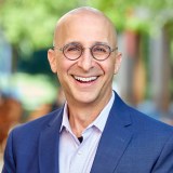 Bald man wearing glasses and a blue suit jacket smiles outdoors with a blurred green background.