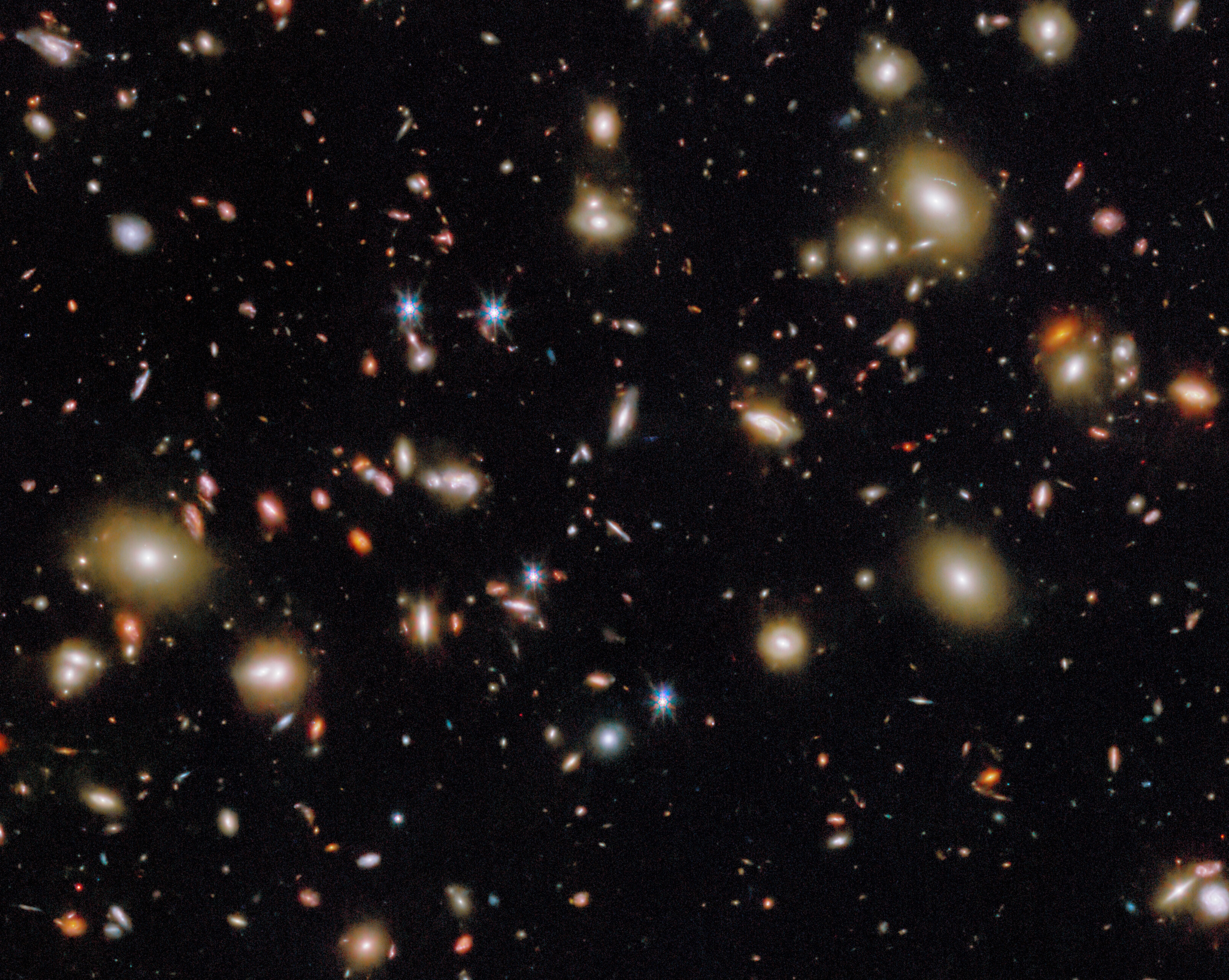 A dense field of galaxies and stars is scattered across a dark background in deep space, with varying sizes, colors, and brightness visible throughout the image.