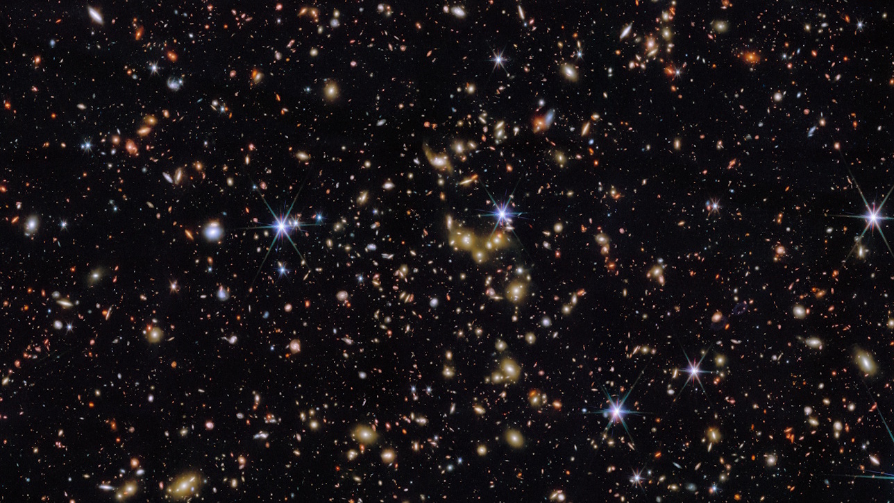 A dense field of stars and galaxies is visible against a black background, with some bright stars showing lens flares.