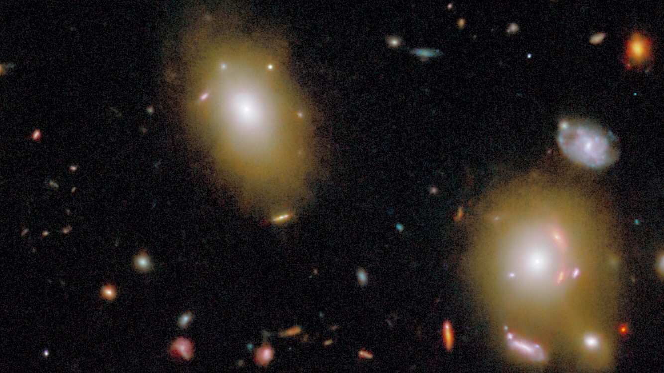 Image of two large elliptical galaxies surrounded by several smaller, colorful galaxies and stars against a dark background in space.