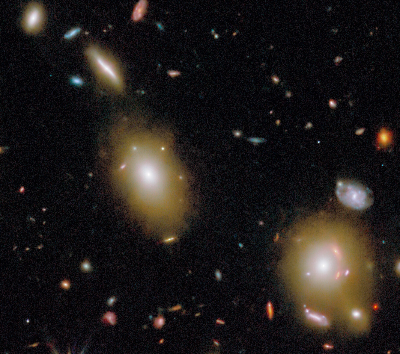 Image of two large elliptical galaxies surrounded by several smaller, colorful galaxies and stars against a dark background in space.
