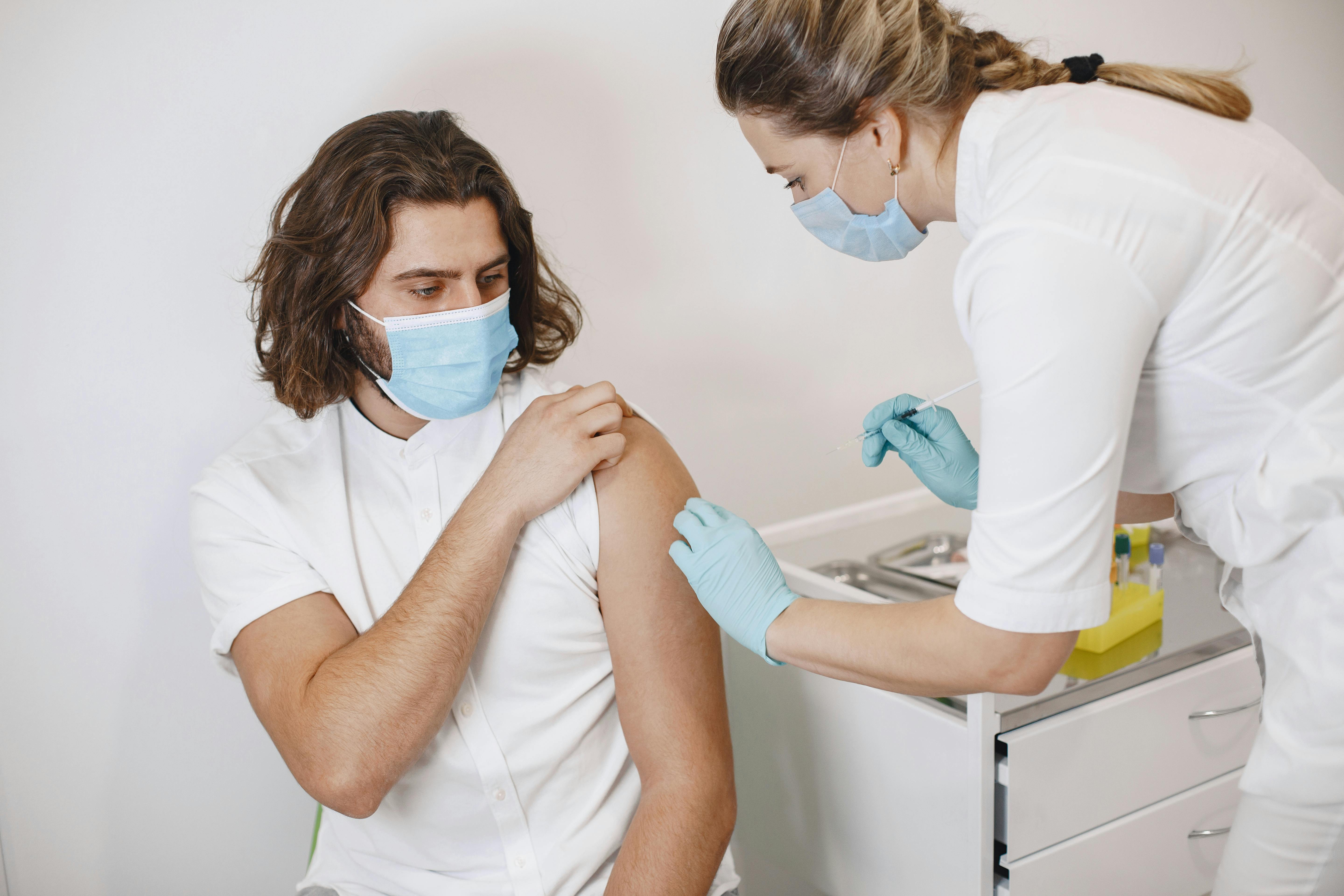 A healthcare worker in gloves and a mask administers a vaccine injection into a seated man's upper arm. Both are wearing face masks, highlighting the ongoing importance of vaccines&mdash;do your own research to stay informed.