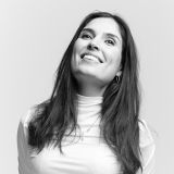 A woman with long dark hair wearing a light turtleneck top smiles and looks upward against a plain background.