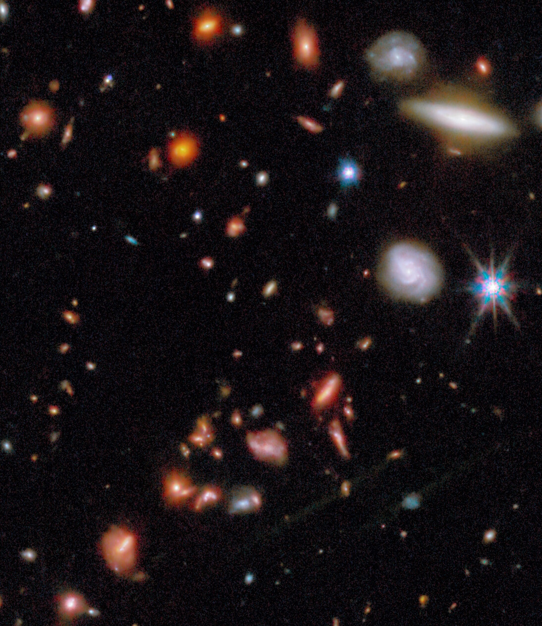 A field of distant galaxies in deep space with various shapes and colors, set against a dark background scattered with faint stars.