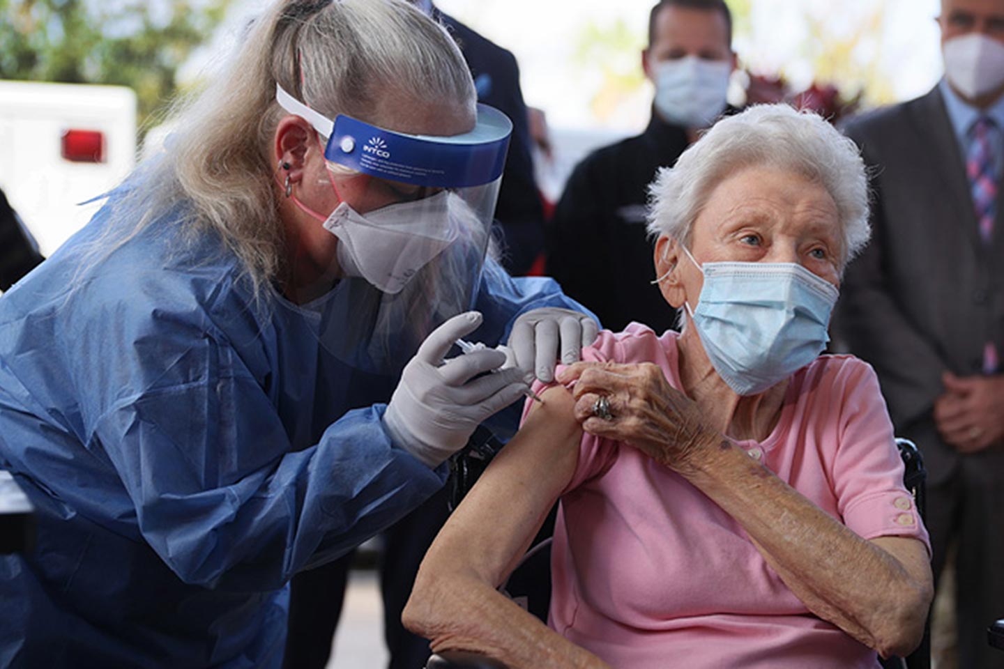 A healthcare worker in protective gear administers a vaccine to an elderly woman wearing a mask, while others in masks stand in the background—highlighting the importance of vaccines and encouraging everyone to do your own research.