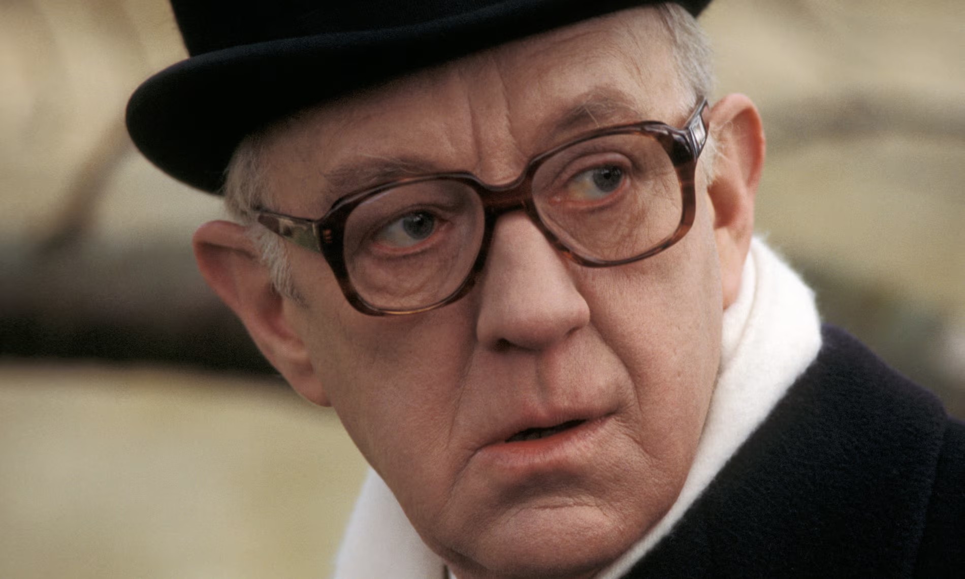 An older man wearing large glasses, a black hat, and a white scarf looks to the side with a serious expression.