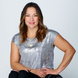 A woman with long brown hair, wearing a silver top and black pants, sits and smiles in a manual wheelchair against a plain white background.