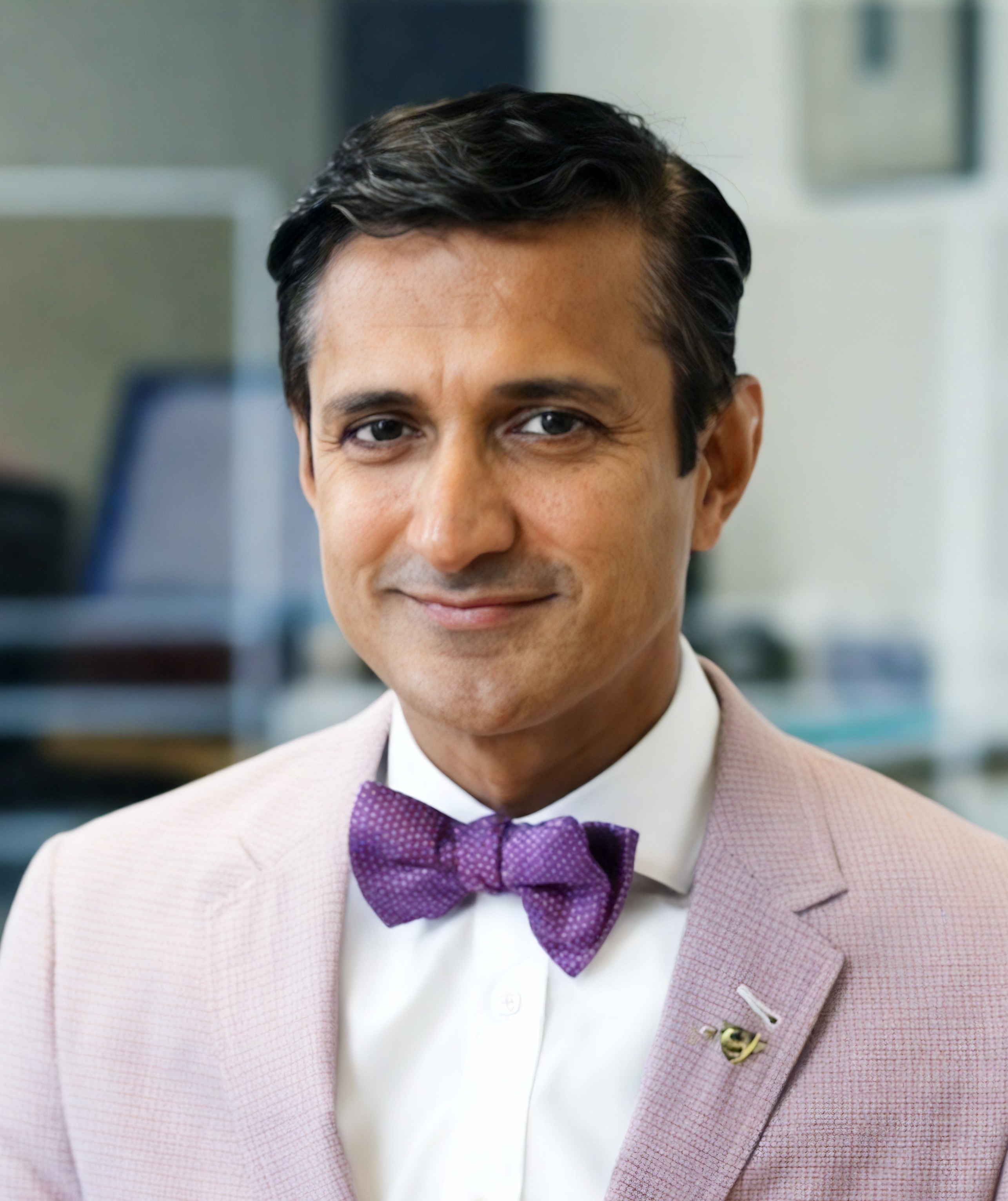 A man wearing a light pink blazer, white shirt, and purple bow tie is smiling slightly while facing the camera.