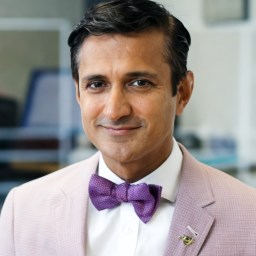 A man wearing a light pink blazer, white shirt, and purple bow tie is smiling slightly while facing the camera.