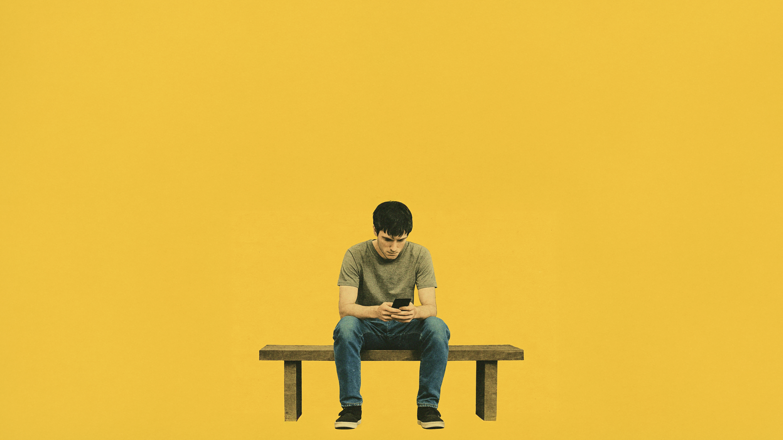 A person sits alone on a bench against a yellow background, looking down at a smartphone.