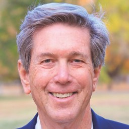 An older man with gray hair and a navy blazer smiles outdoors with blurred trees in the background.