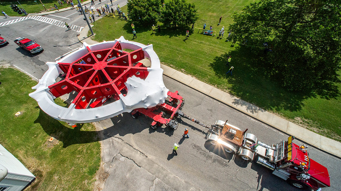 Aerial view of a large red and white amusement park ride section, reminiscent of the muon g-2 anomaly ring, being transported by a semi-truck on a road, with workers nearby and people observing from a grassy area.