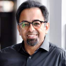 A man with glasses and a goatee, wearing a black shirt, smiles while standing indoors near a window.