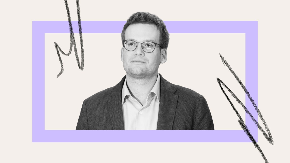 A man in glasses and a suit jacket, resembling John Green, stands in front of a light background with a purple rectangle and abstract black lines.