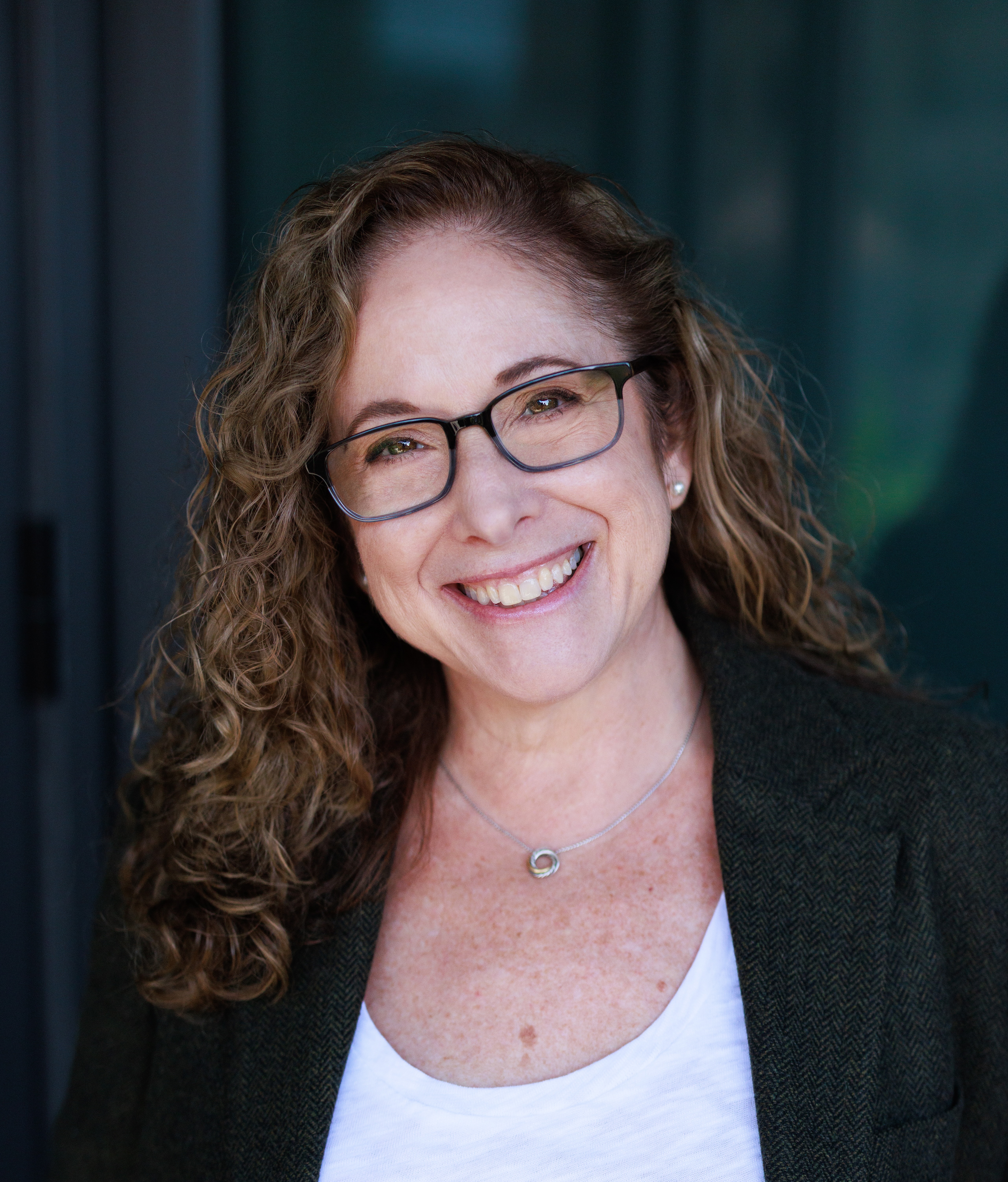 A woman with curly hair and glasses smiles at the camera. She is wearing a dark blazer over a white top and a necklace, standing in front of a dark, blurred background.
