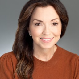 Woman with long brown hair wearing a rust-colored top, smiling at the camera against a plain gray background.