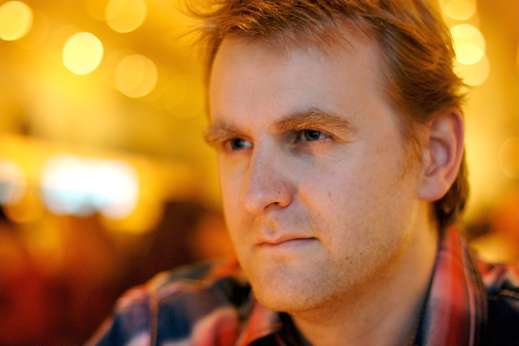 A man with light brown hair and a slight beard is looking thoughtfully ahead, with warm, out-of-focus lights in the background.