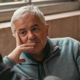 An older man with gray hair, wearing a grey zip-up sweater, rests his chin on his hand while sitting indoors near a window.