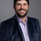 Man with short dark hair and beard, wearing a blue velvet jacket over a light purple shirt, smiles in front of a plain gray background.