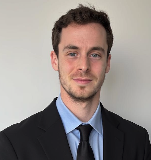 A man with short brown hair and light facial hair wearing a black suit, light blue shirt, and black tie, standing in front of a plain light-colored wall.