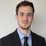 A man with short brown hair and light facial hair wearing a black suit, light blue shirt, and black tie, standing in front of a plain light-colored wall.