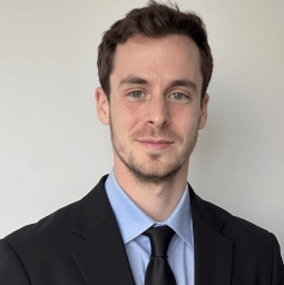 A man with short brown hair and light facial hair wearing a black suit, light blue shirt, and black tie, standing in front of a plain light-colored wall.