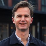 A man with light brown hair, wearing a dark jacket over a light shirt, stands outdoors and smiles at the camera.