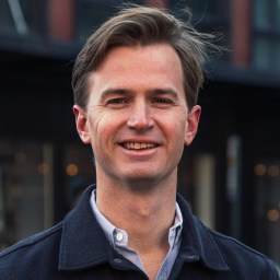 A man with light brown hair, wearing a dark jacket over a light shirt, stands outdoors and smiles at the camera.