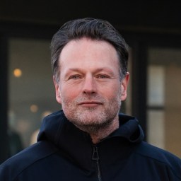 A middle-aged man with short dark hair and a trimmed beard, wearing a black jacket, stands outdoors in front of a blurred building background.