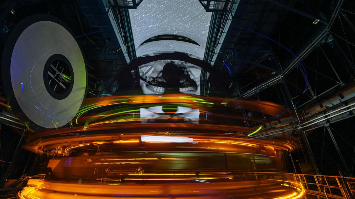 Interior view of a large observatory telescope in operation at night, with orange light trails and a starry sky visible through the open roof.
