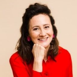 Woman with dark hair wearing a red blouse and black pants sits on a stool, smiling at the camera with her chin resting on her hand against a beige background.