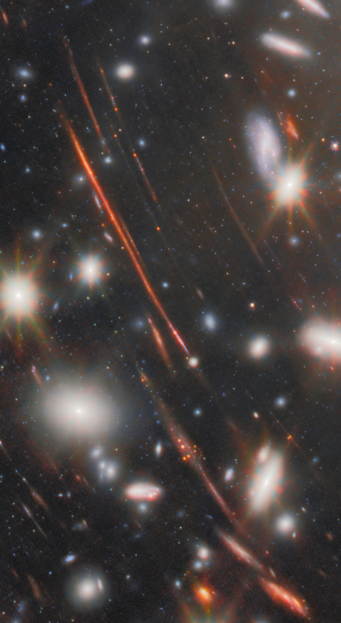 A deep-space image from JWST shows bright galaxies, numerous stars, and several long, red streaks created by the most extreme gravitational lens, all set against a dark background filled with distant celestial objects.