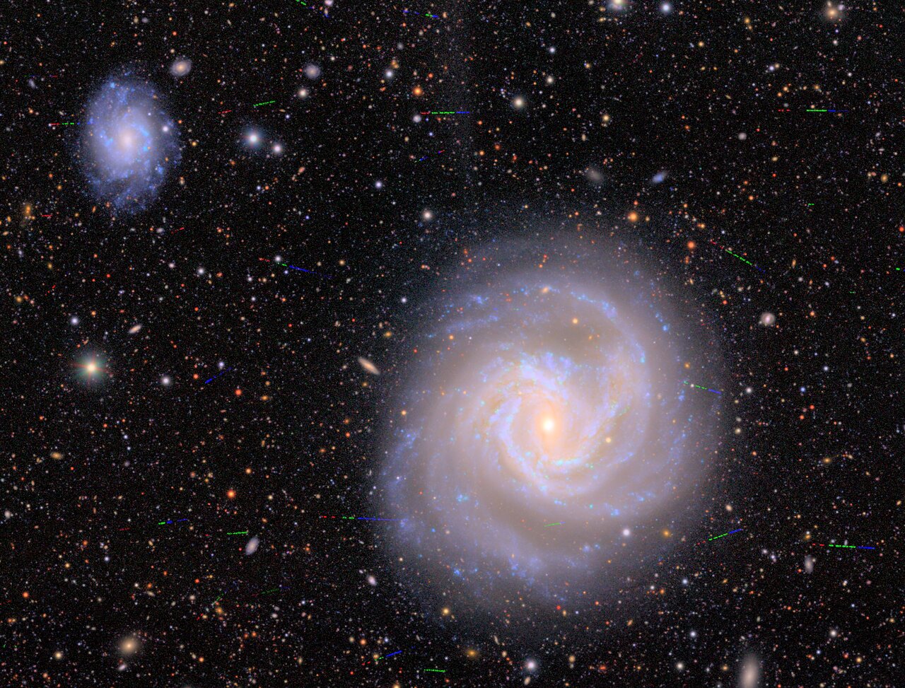 Spiral galaxy with a bright core and swirling arms is centered, as captured by the Vera Rubin Observatory, with another smaller galaxy in the upper left, surrounded by stars and distant galaxies that invite astronomers to solve cosmic puzzles.