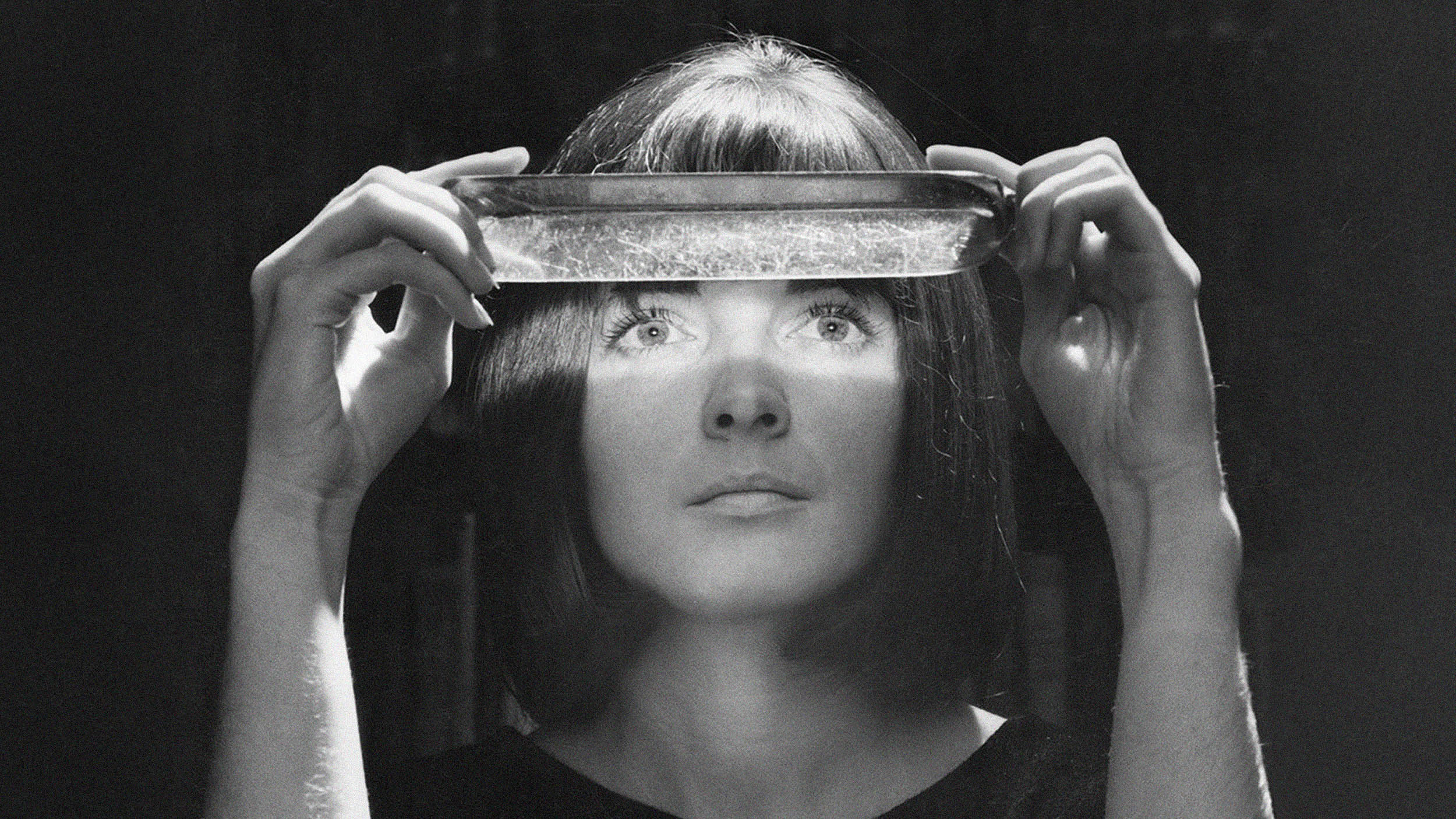 A woman, resembling a paranormal investigator, holds a rectangular glass dish above her eyes, which are illuminated by light shining through the dish in a dark setting.