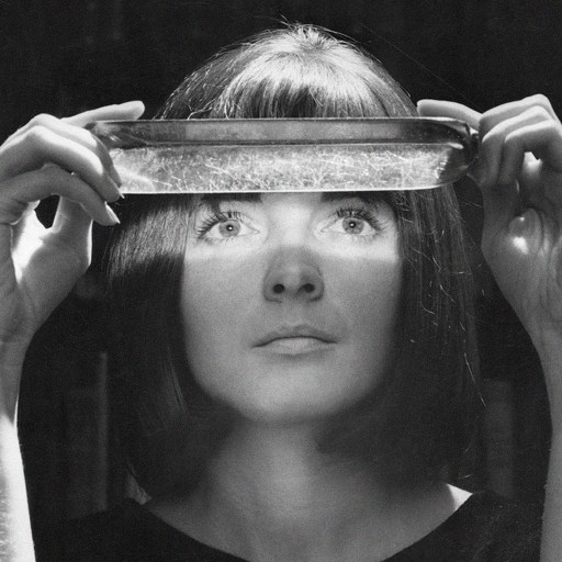 A woman, resembling a paranormal investigator, holds a rectangular glass dish above her eyes, which are illuminated by light shining through the dish in a dark setting.